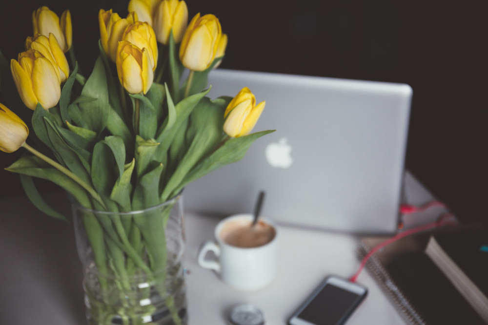 Ein Tisch mit einer Vase voller gelber Tulpen, im Hintergrund sieht man einen Laptop, ein Smartphone und ein Kaffeetasse.